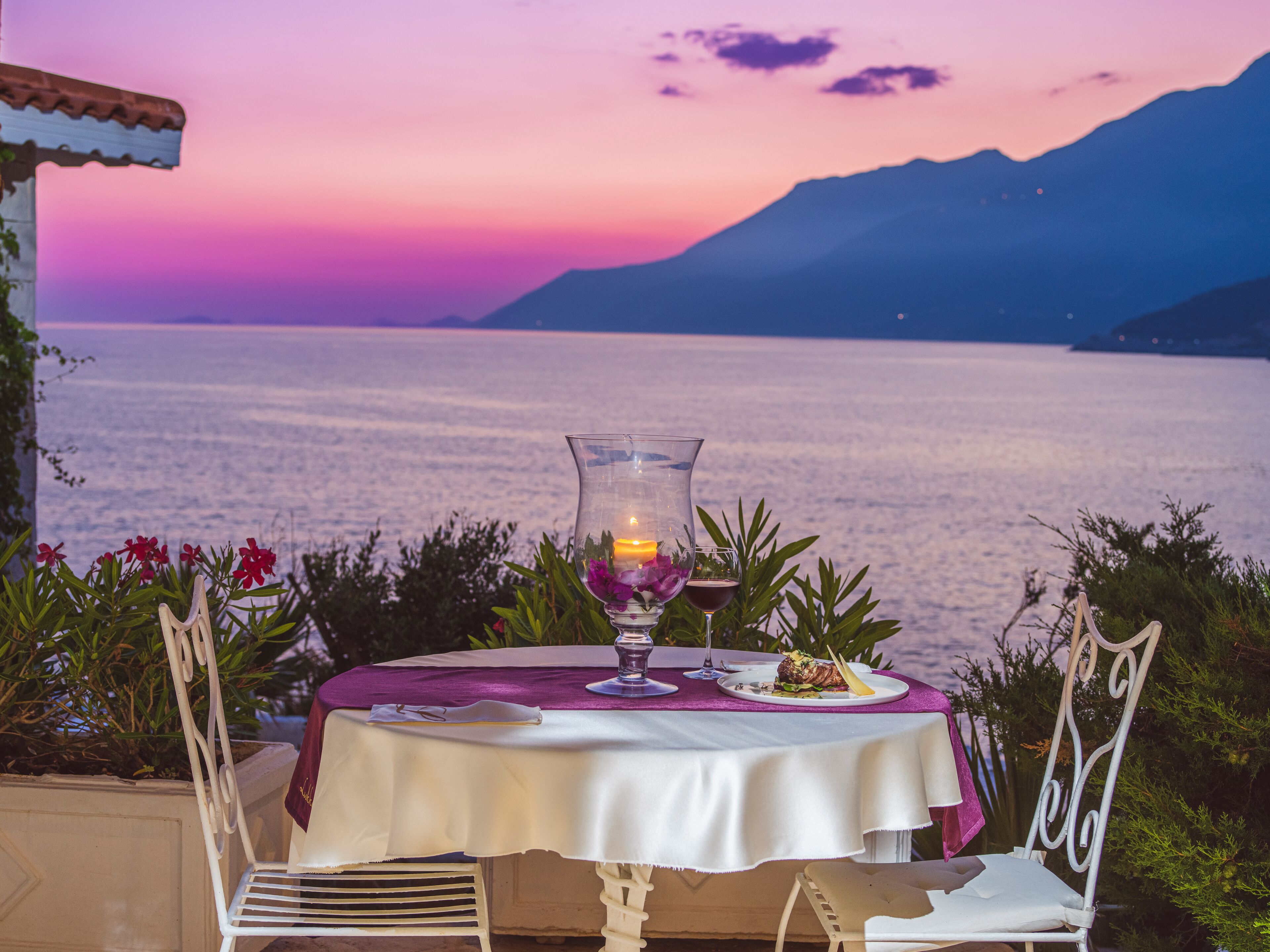 A table set for two with a candle, a plate of food, and a view of the sea at sunset.