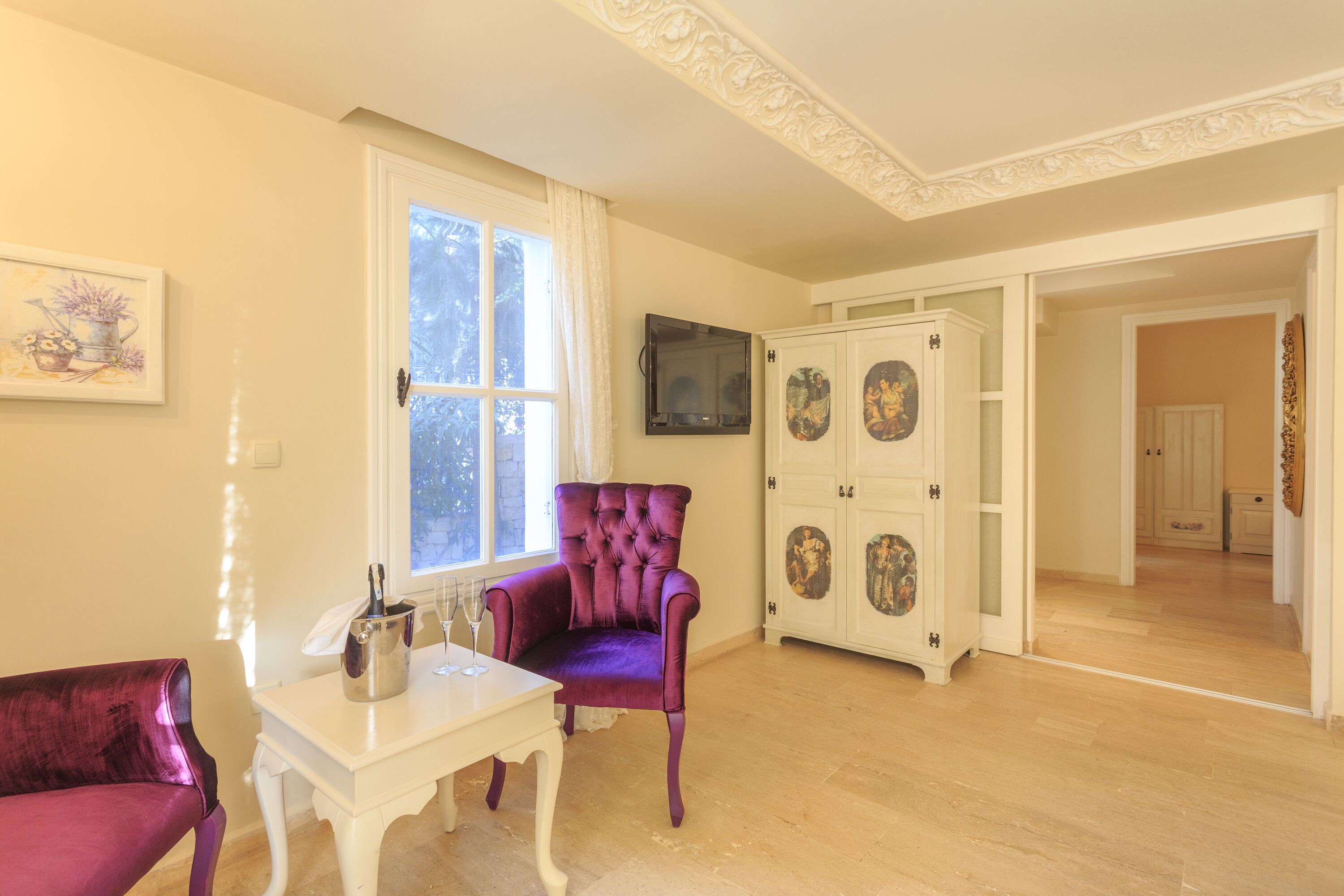 Una habitación con piso de madera, una mesa blanca con un florero y una botella, una silla morada y un armario blanco con puertas decorativas.