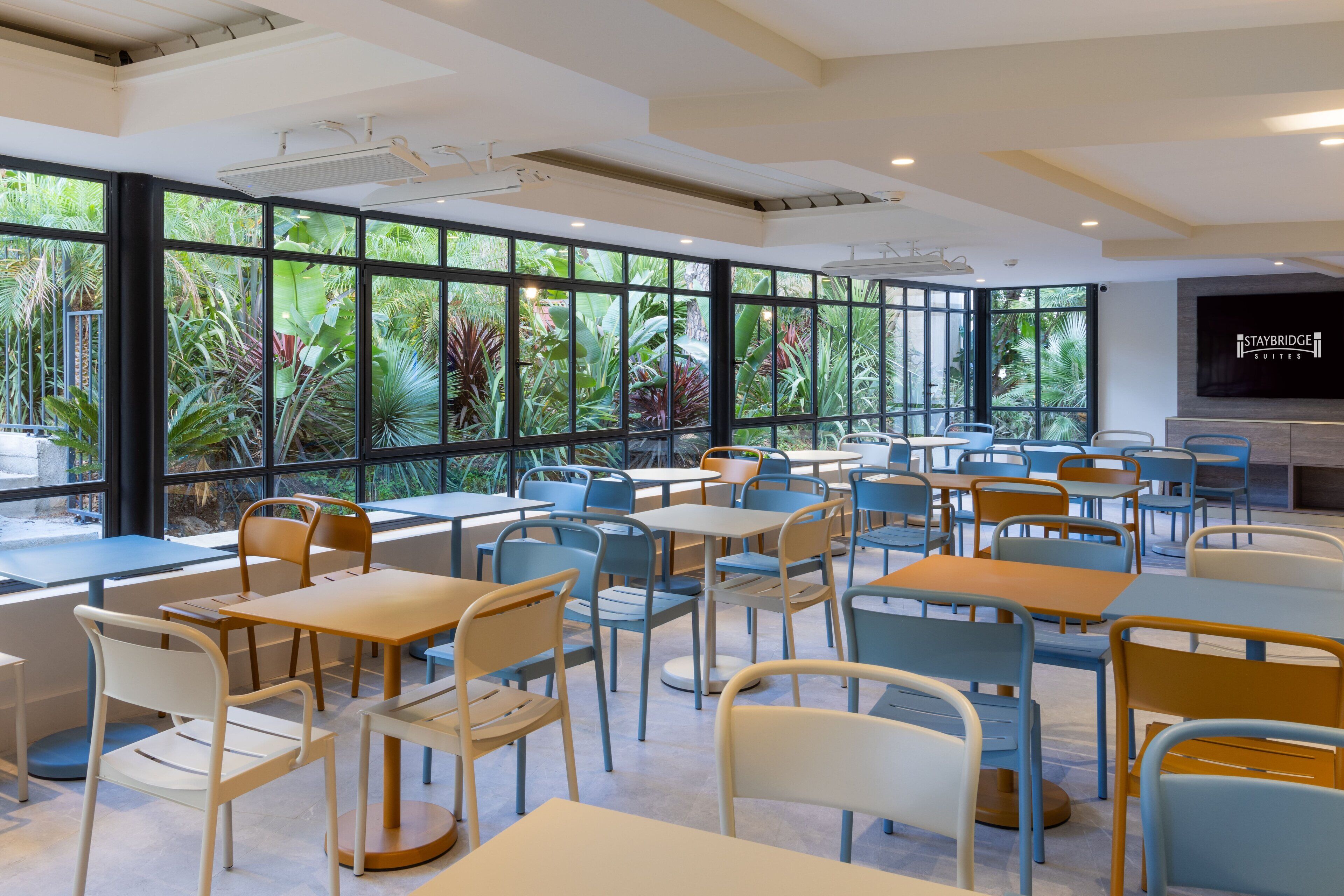Un comedor moderno con mesas y sillas, ventanas amplias con vistas a zonas verdes y una pantalla en la pared mostrando 'Staybridge Suites'.