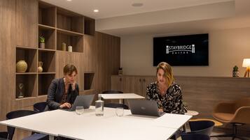 Uma sala de conferências com duas pessoas trabalhando em laptops, uma TV exibindo 'STAYBRIDGE QUINTES' e uma estante de madeira com objetos decorativos.