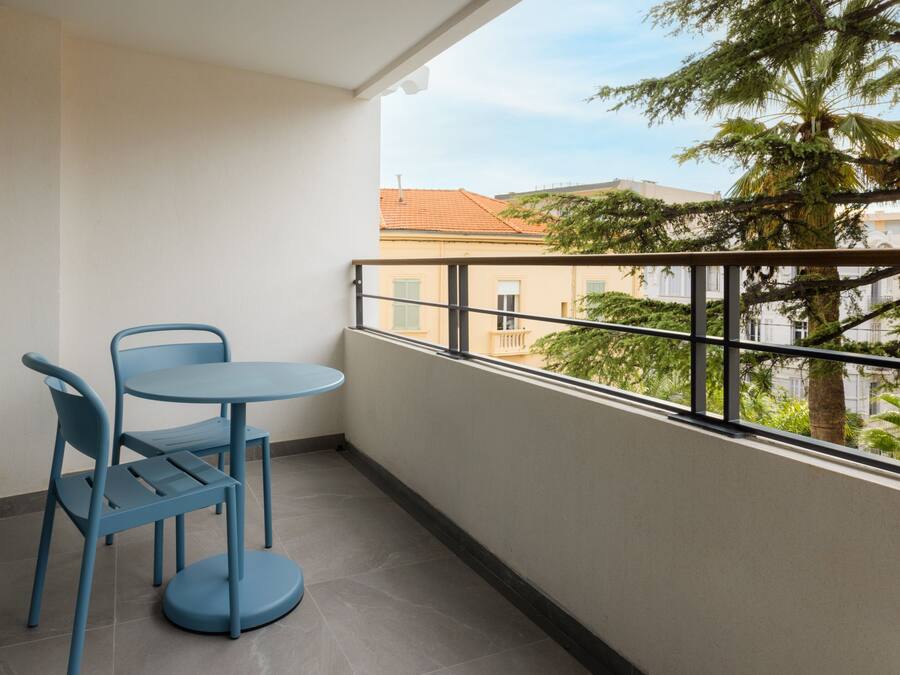 Ein Balkon mit rundem Tisch und zwei Stühlen, Blick auf Gebäude und Bäume, und einem Metallgeländer.
