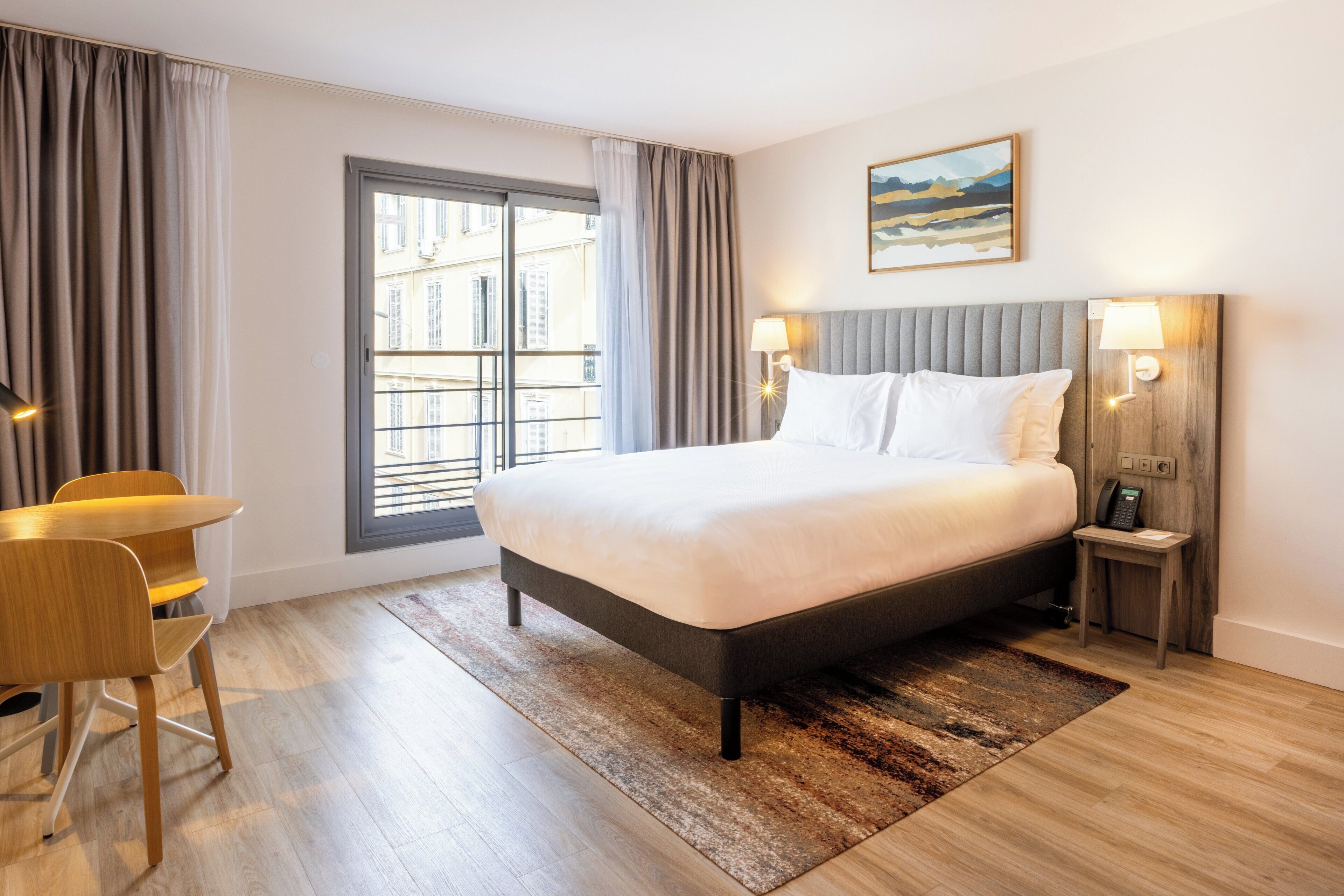 A hotel room with a large bed, a desk, and a chair. There is a window with curtains, a nightstand with a lamp, and a painting on the wall.
