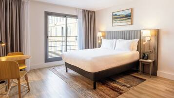 A hotel room with a large bed, a desk, and a chair. There is a window with curtains, a nightstand with a lamp, and a painting on the wall.
