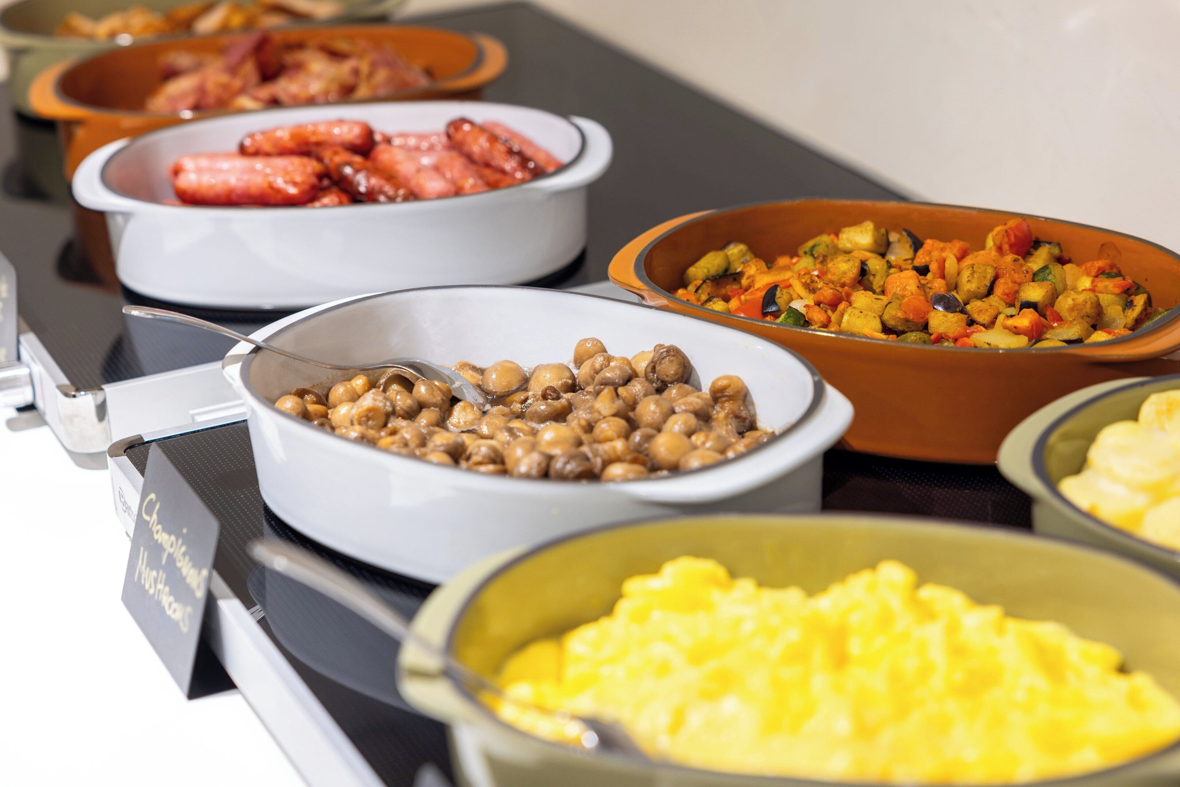 A buffet with various dishes including sausages, chickpeas, and scrambled eggs.