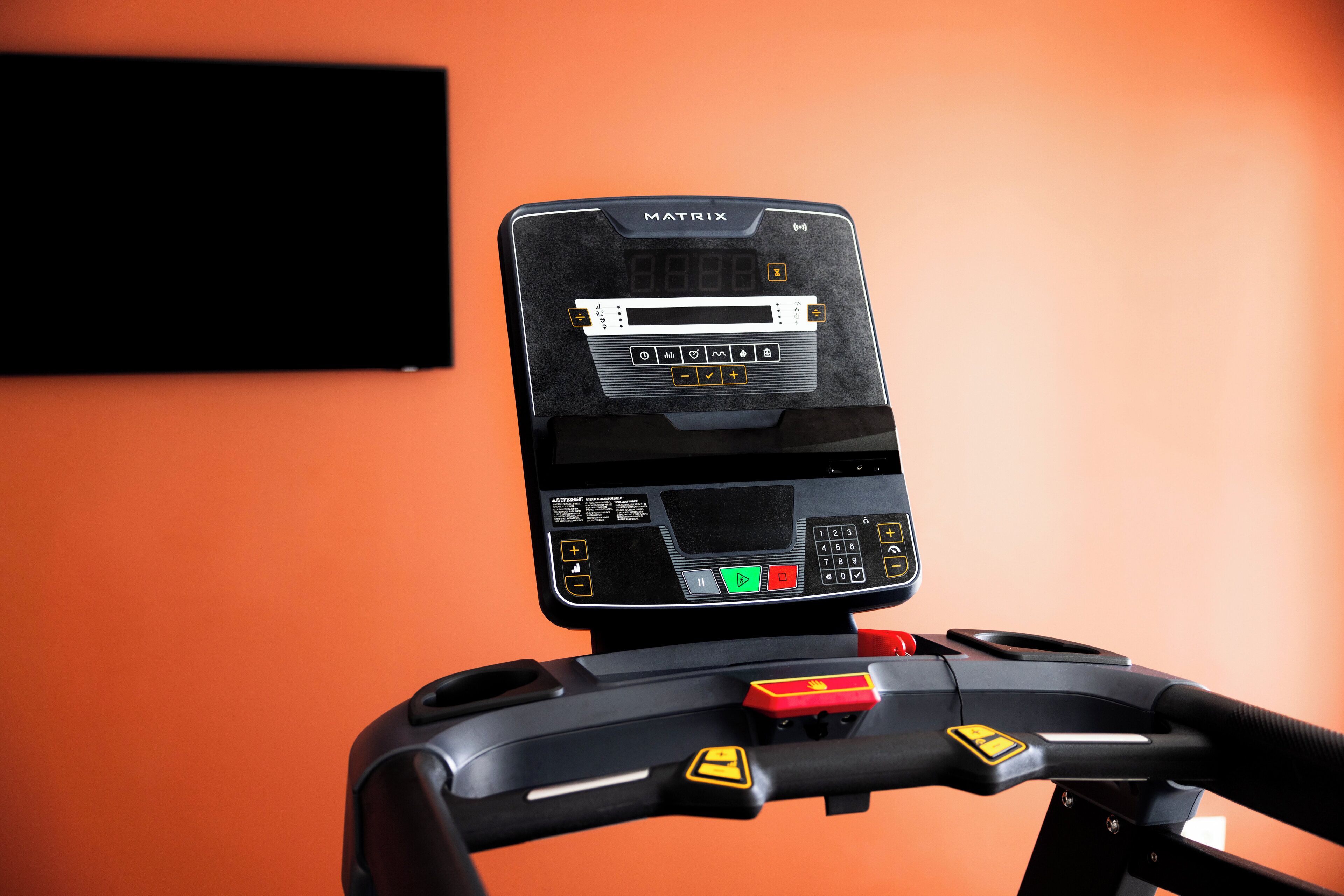 A treadmill with a digital display and control panel, set against an orange wall.