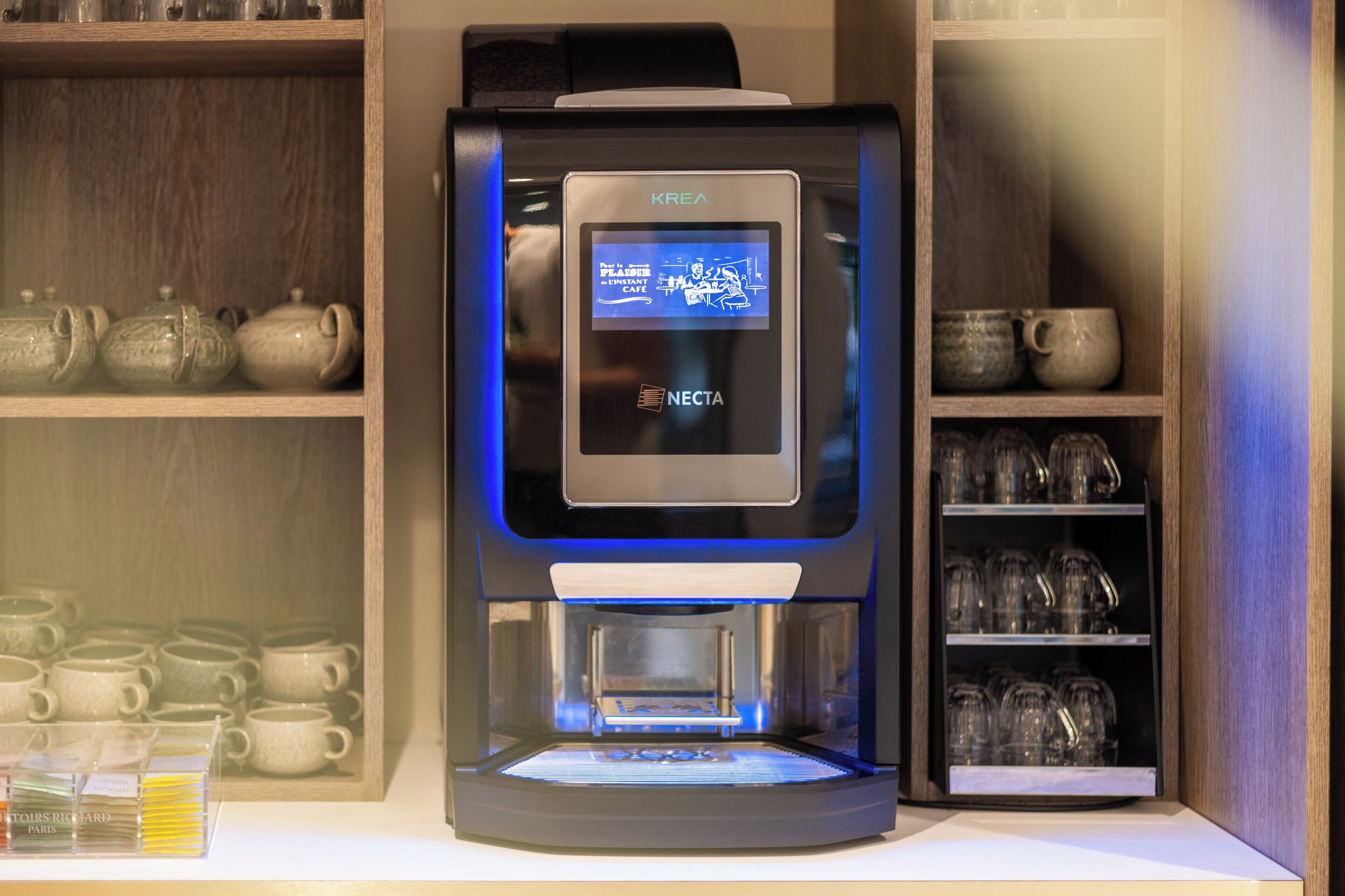 Une machine à café dotée d’un écran numérique, entourée d’étagères où sont disposées des tasses et des théières en céramique.