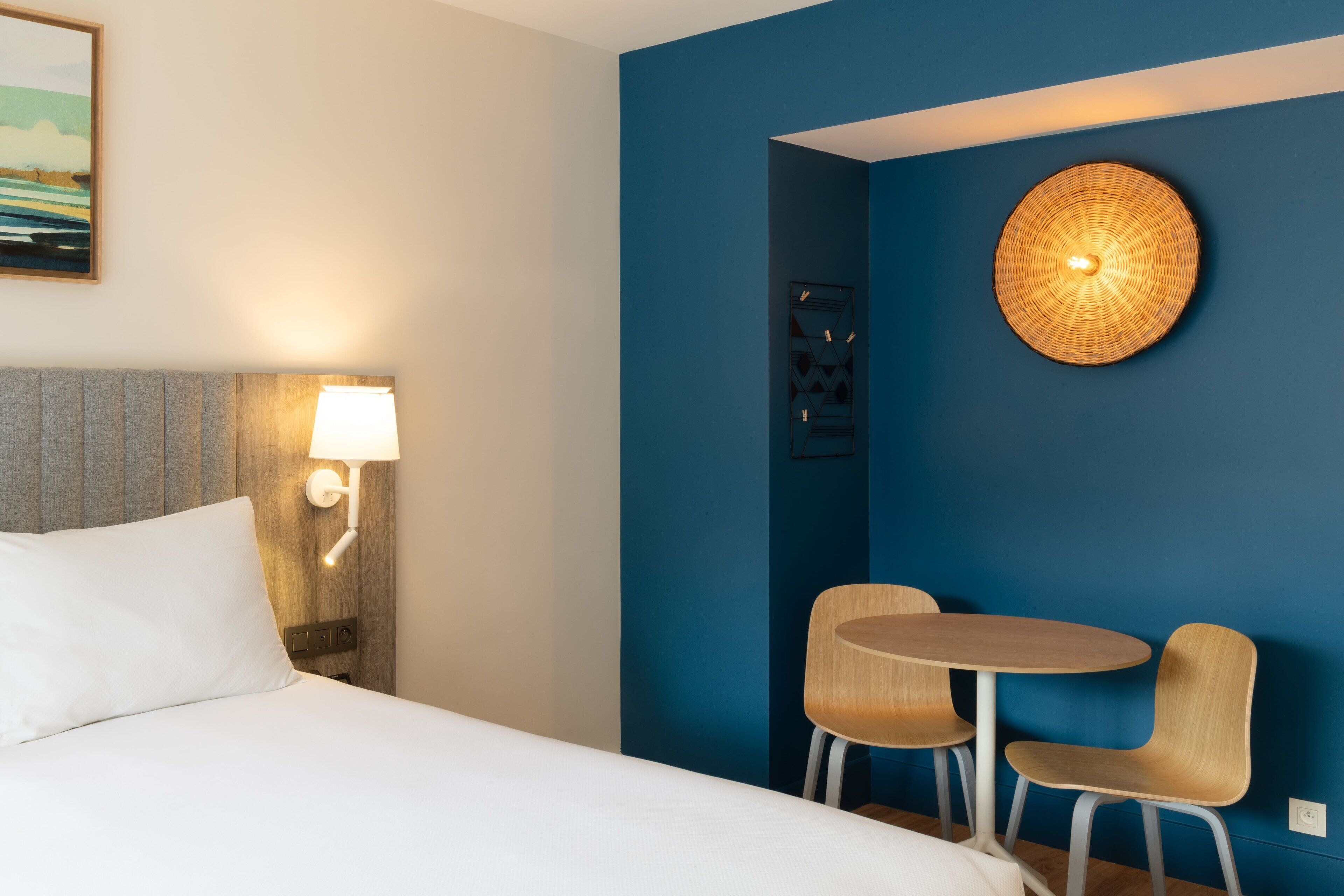 A hotel room with a bed, a nightstand with a lamp, a round wall hanging, and two chairs around a small table.