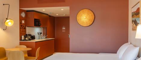 A hotel room with two beds, a small kitchenette, a dining area with chairs, and a wall-mounted lamp.