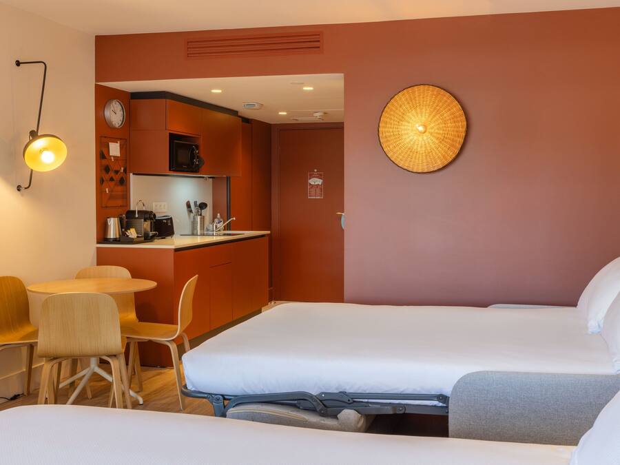 Habitación de hotel con dos camas, una kitchenette pequeña, un área de comedor con sillas y una lámpara fijada a la pared.