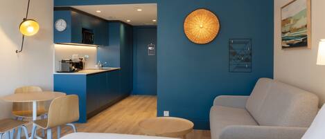 A modern hotel room with a kitchenette, a dining area with a round table and chairs, a sofa, and a large wall-mounted lamp.