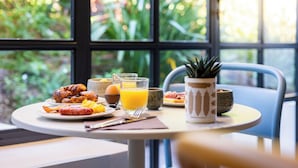 Una mesa de desayuno con croissants, huevos y zumo de naranja, frente a una ventana con vistas a la vegetación.