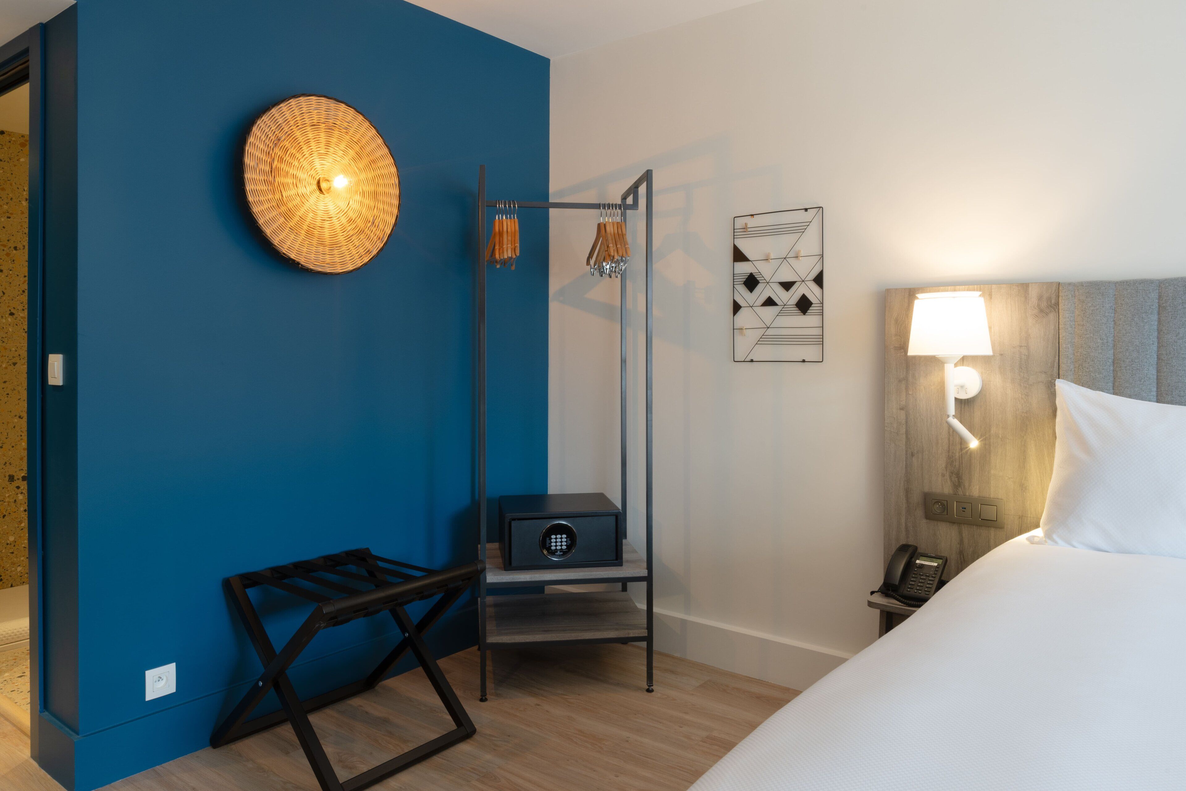 Une chambre d’hôtel avec un mur d’accent bleu, un lit avec une literie blanche, une table de chevet avec une lampe, un téléphone et une petite armoire.