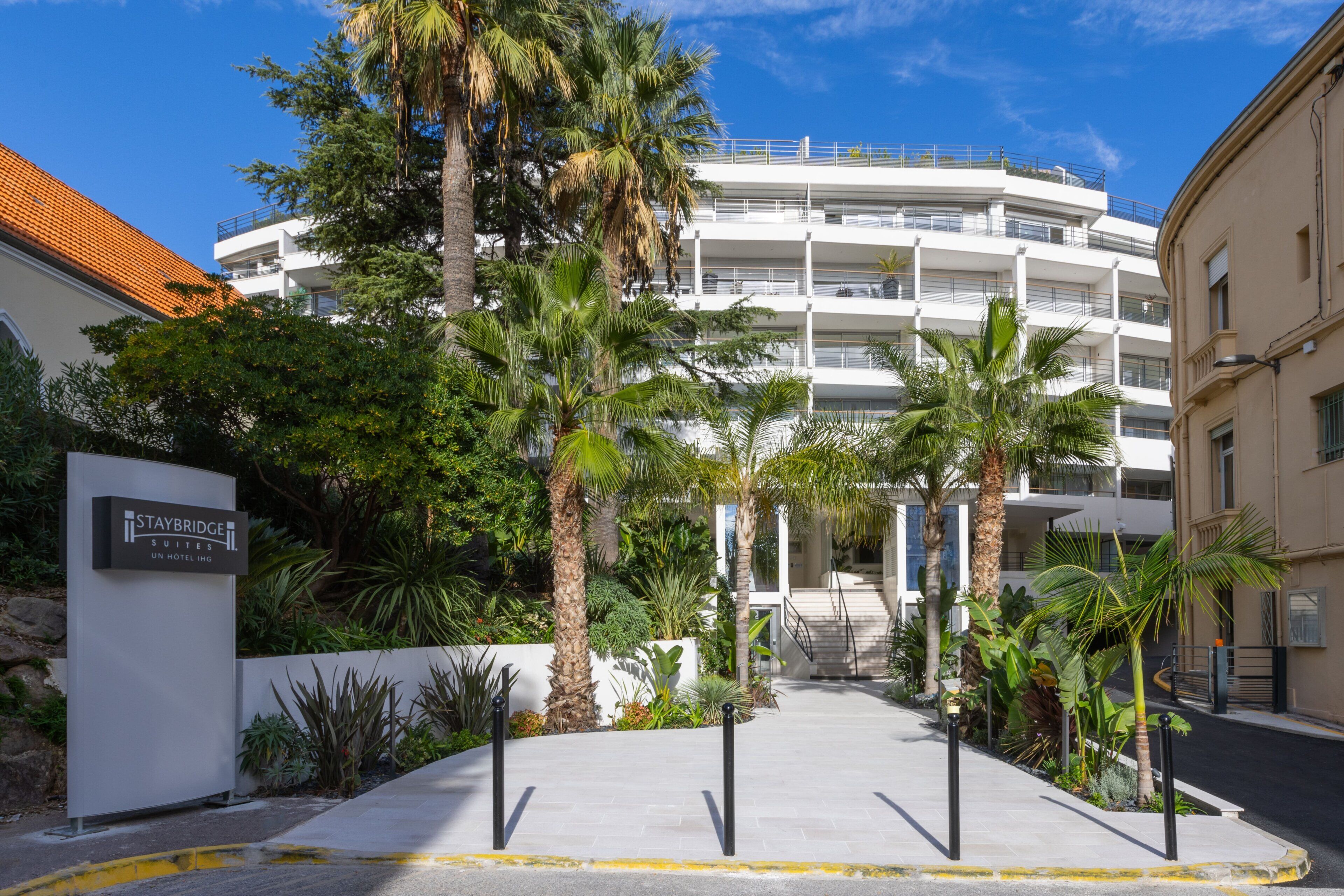 A hotel entrance with a sign for Staybridge Suites, a palm tree-lined pathway, and a multi-story building in the background.