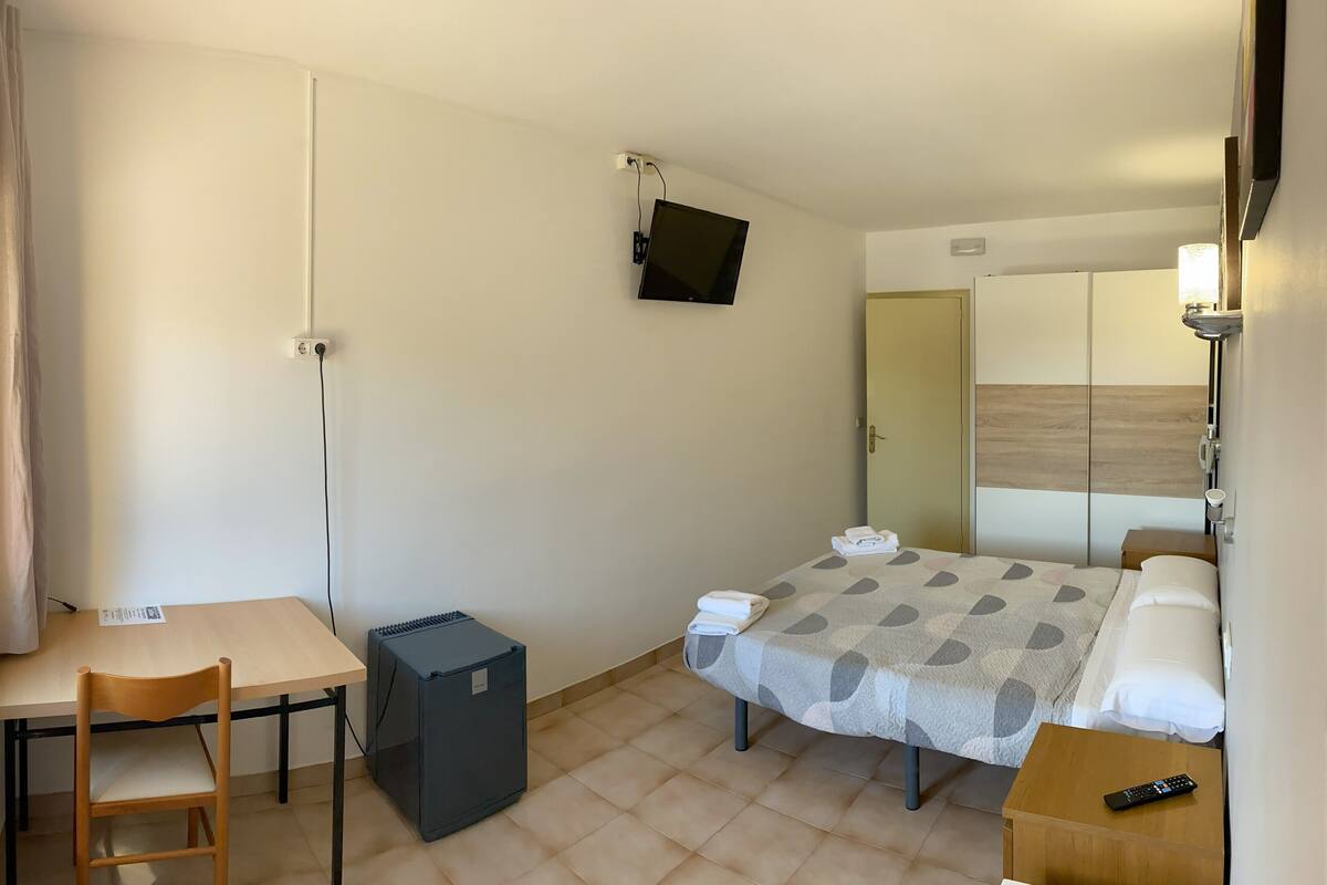A small, tidy room with a bed, a desk, a chair, a small table, a heater, and a TV mounted on the wall.