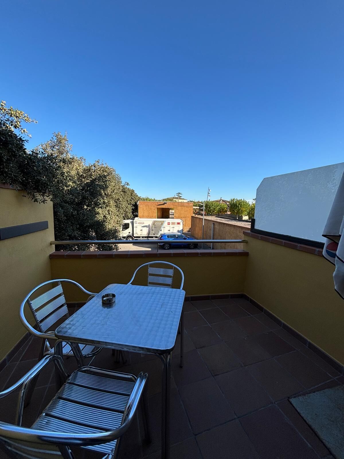 Eine kleine Außenpatio mit einem Tisch, zwei Stühlen, einem Grill und Blick auf die Straße und Gebäude im Hintergrund.