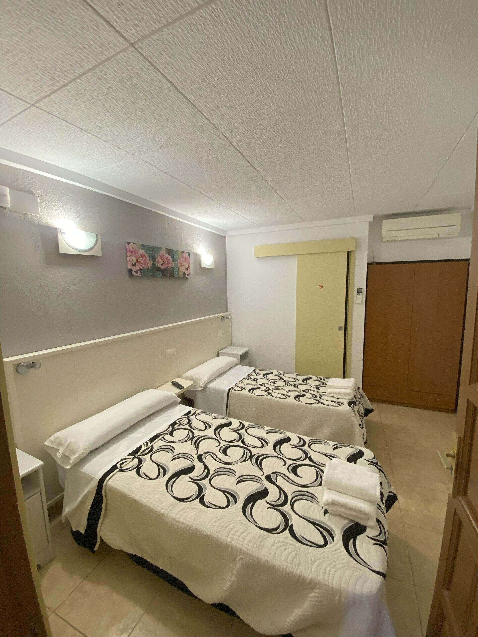 A hotel room with two beds, a small table, a wall-mounted light, and a door.