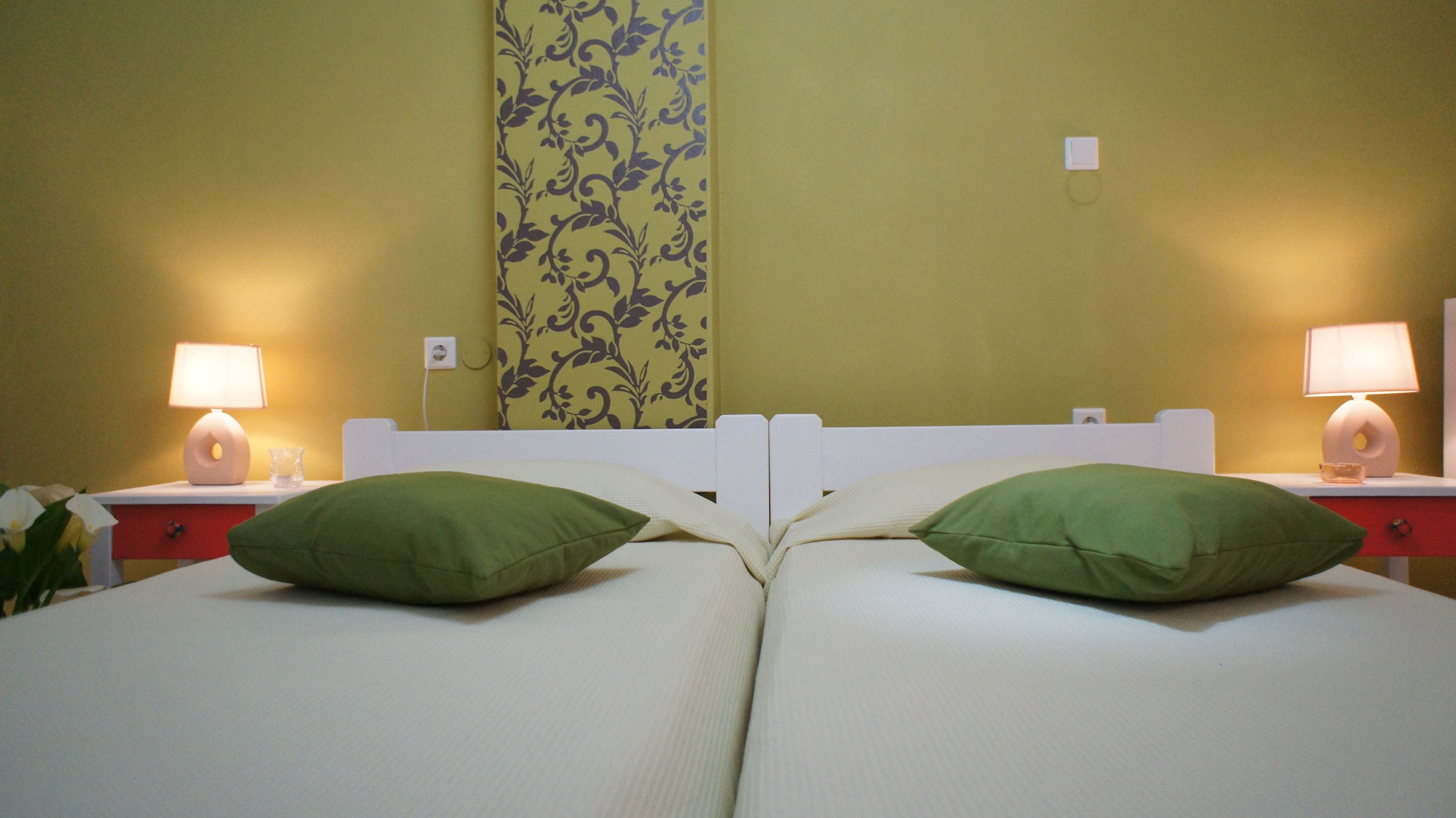 A double bed with green pillows, a nightstand with a lamp, and a floral-patterned wall.