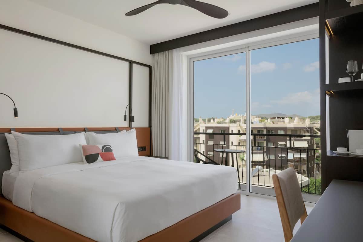 A hotel room with a large bed, a desk, and a balcony with a city view.