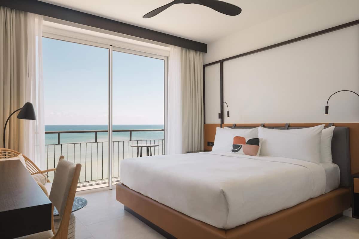 A hotel room with a large bed, a desk, and a chair. There is a balcony with a view of the ocean.
