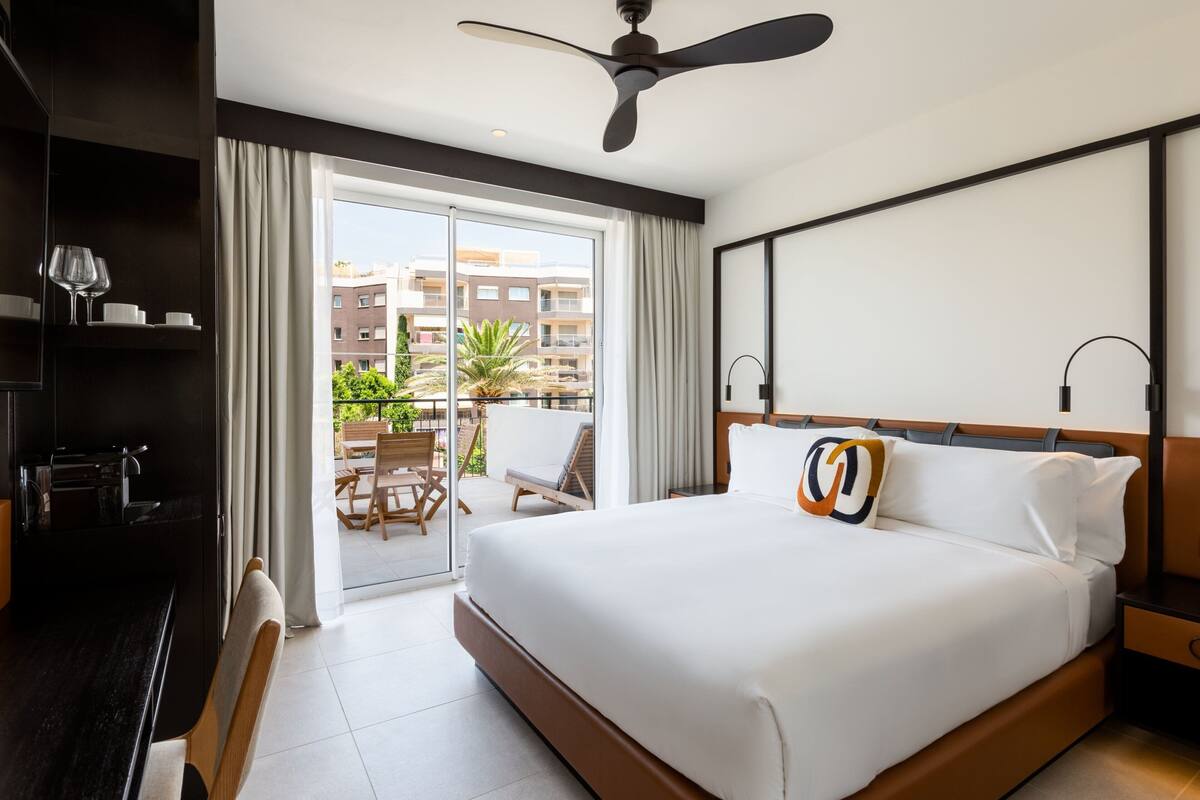 A hotel room with a large bed, a ceiling fan, a balcony with a view of the city, and a dining area with chairs and a table.