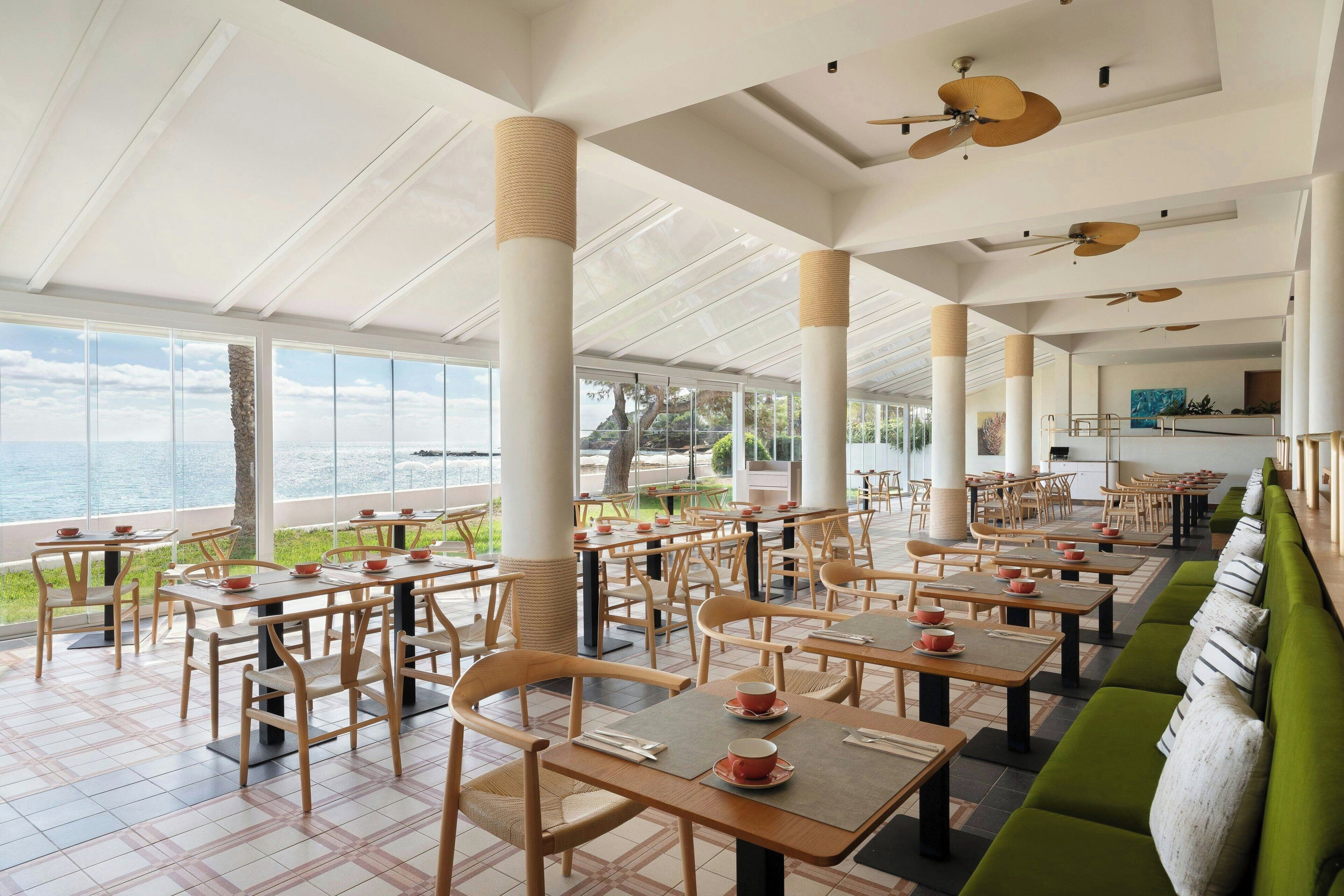 Un amplio espacio exterior con zona de comedor, varias mesas y sillas, ventanas grandes con vistas al mar y ventiladores de techo.