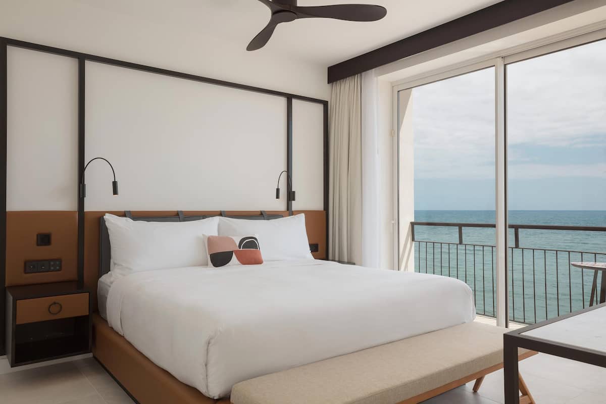 A hotel room with a large bed, a bedside table, a ceiling fan, a window with a view of the sea, and a balcony with a table.
