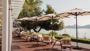An outdoor seating area with wooden chairs and tables, large umbrellas, and a view of the water. - Hotel Riomar Ibiza, A Tribute Portfolio Hotel (Santa Eulalia del Rio)
