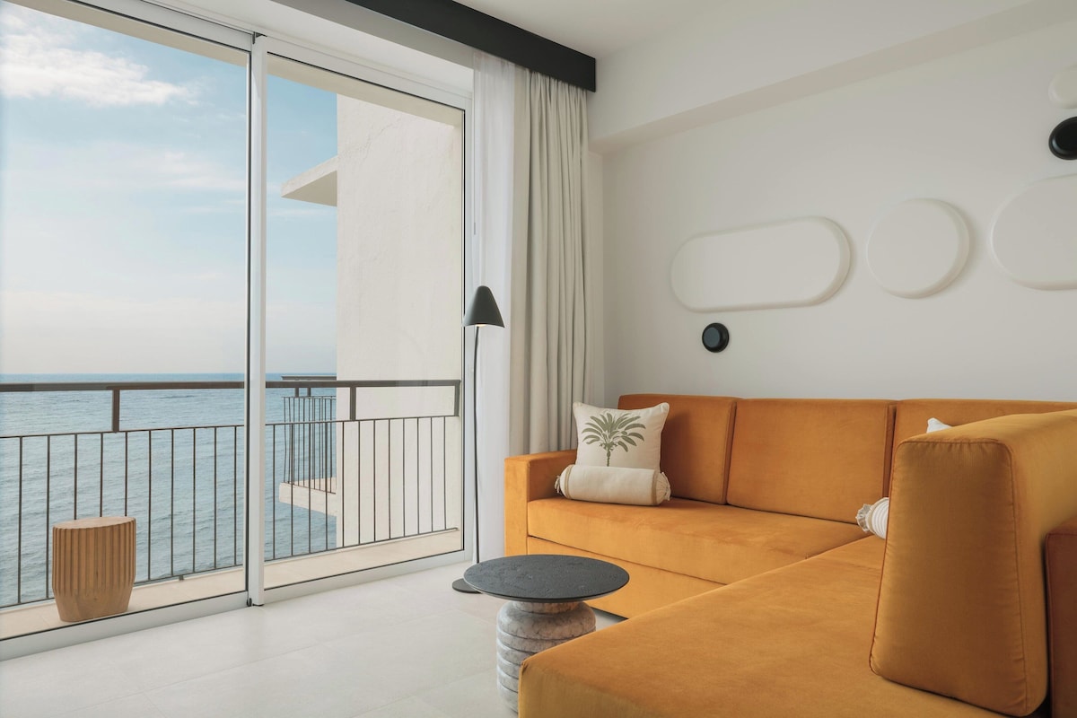 Un salon moderne avec un canapé, une table basse ronde et une vue sur la mer, visible à travers une porte-fenêtre coulissante.