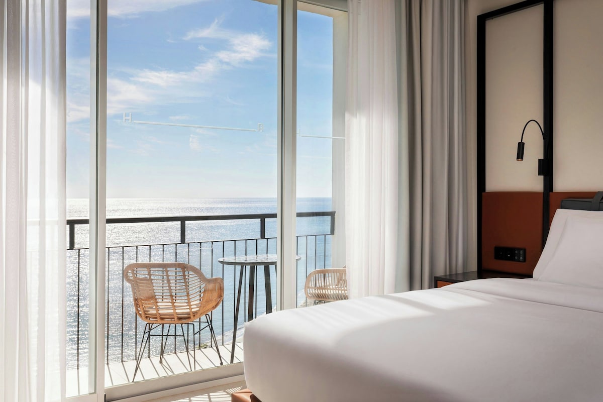 Une chambre d’hôtel avec un grand balcon, un lit, deux chaises et une table de chevet.