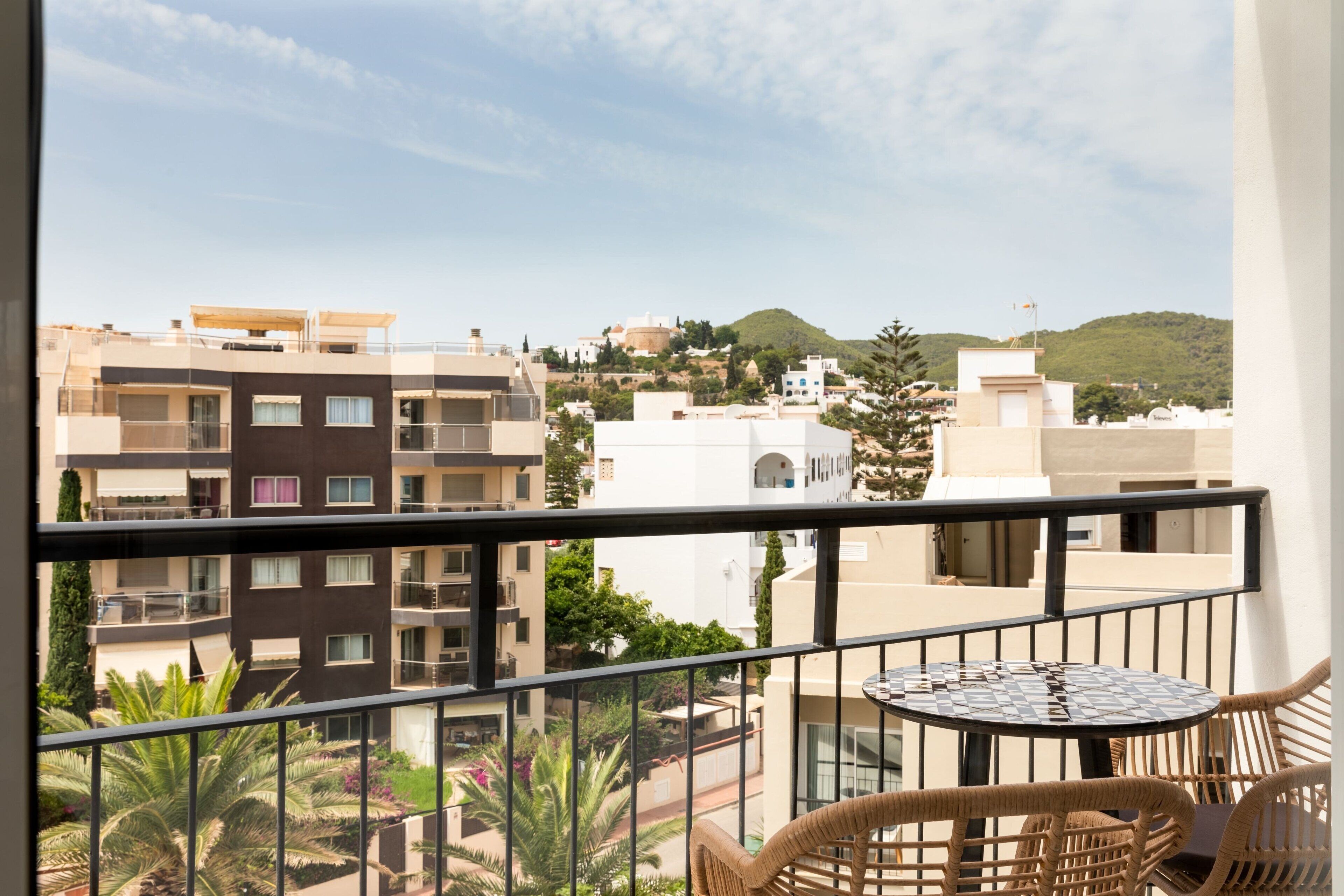 Ein Balkon mit Korbmöbeln, der auf einen Stadtblick mit verschiedenen Gebäuden und einer Hügelkulisse im Hintergrund blickt.