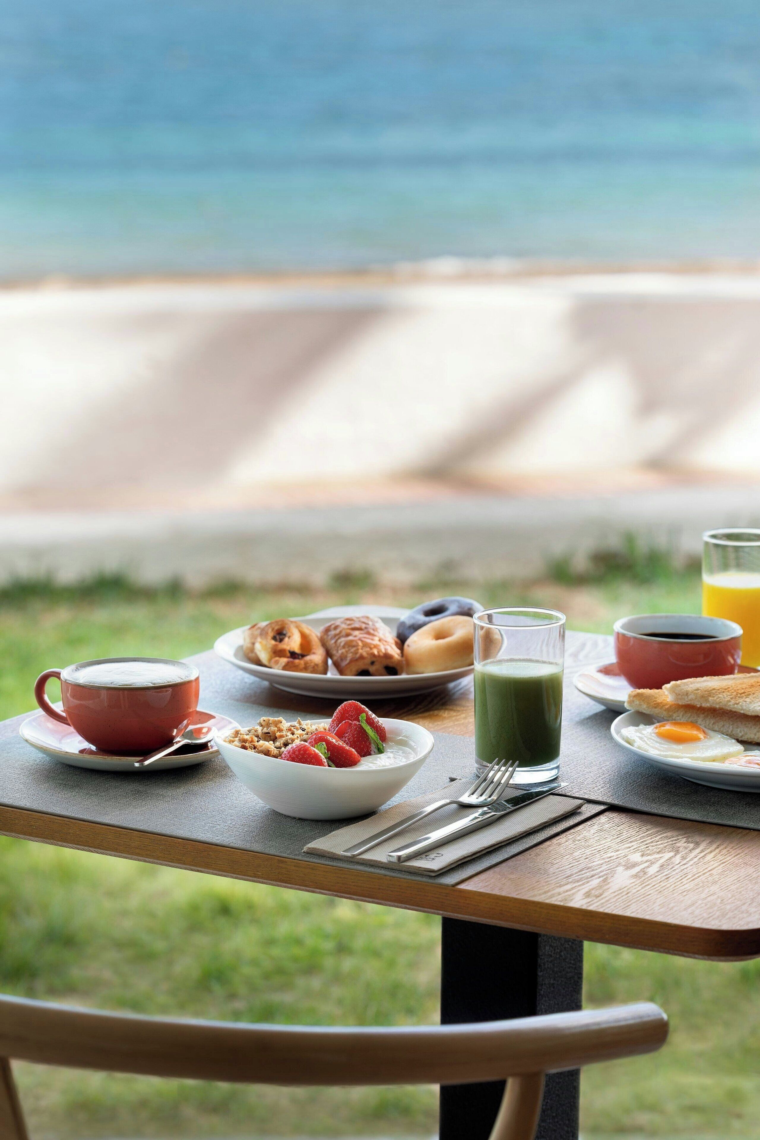 A breakfast table with various food items and a glass of orange juice, overlooking a beach.