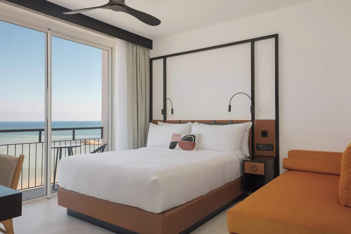 A hotel room with a large bed, a nightstand, an orange sofa, and a balcony with a view of the ocean.