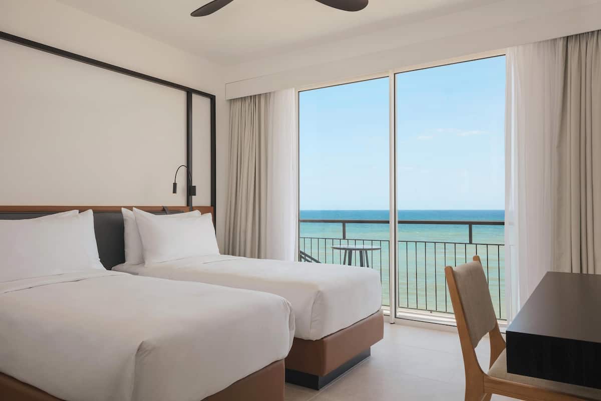 A hotel room with two beds, a dining table, and a balcony with a view of the ocean.