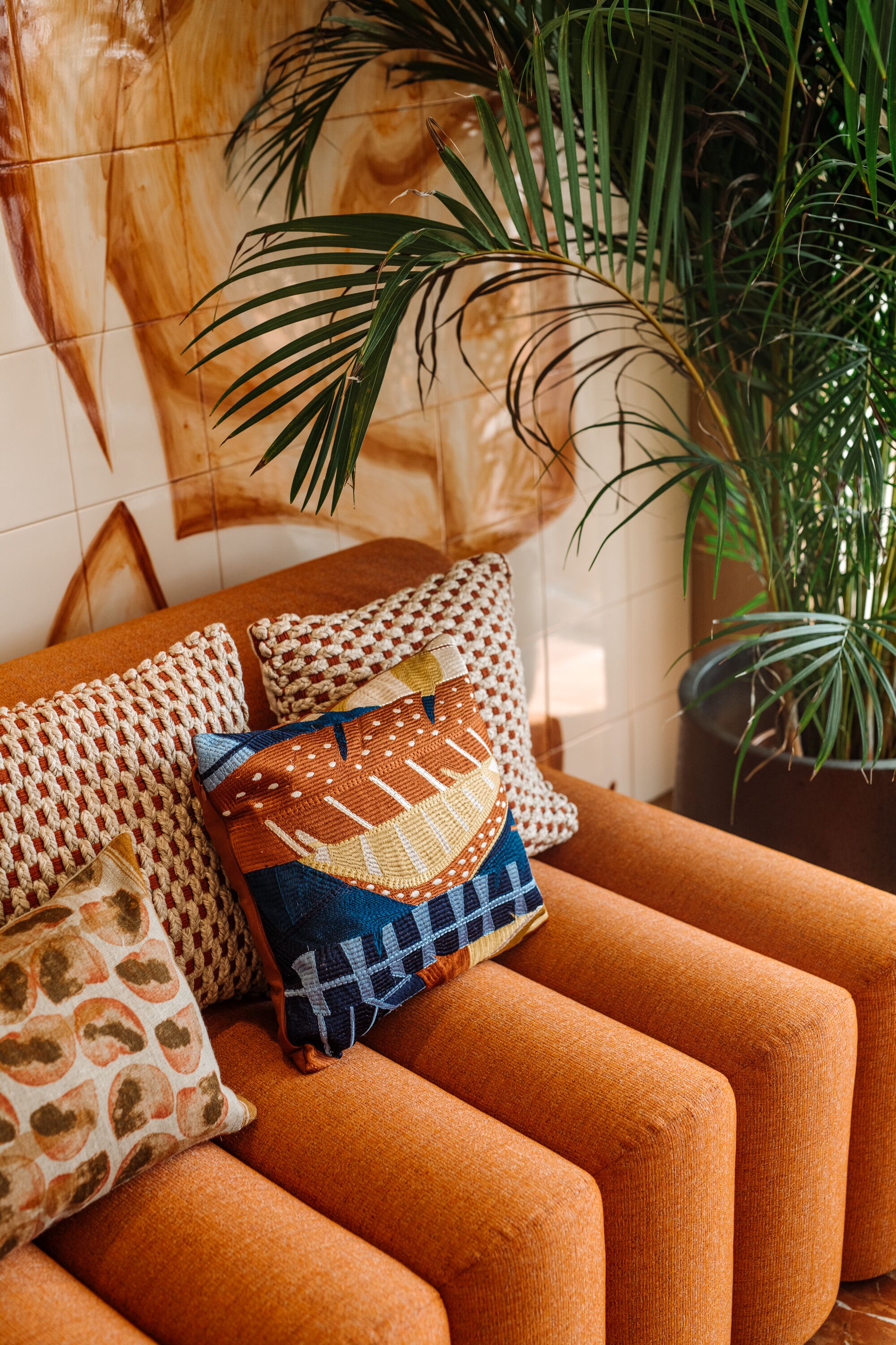 A room with a sofa adorned with patterned cushions, a large potted plant, and a tiled wall with a geometric design.