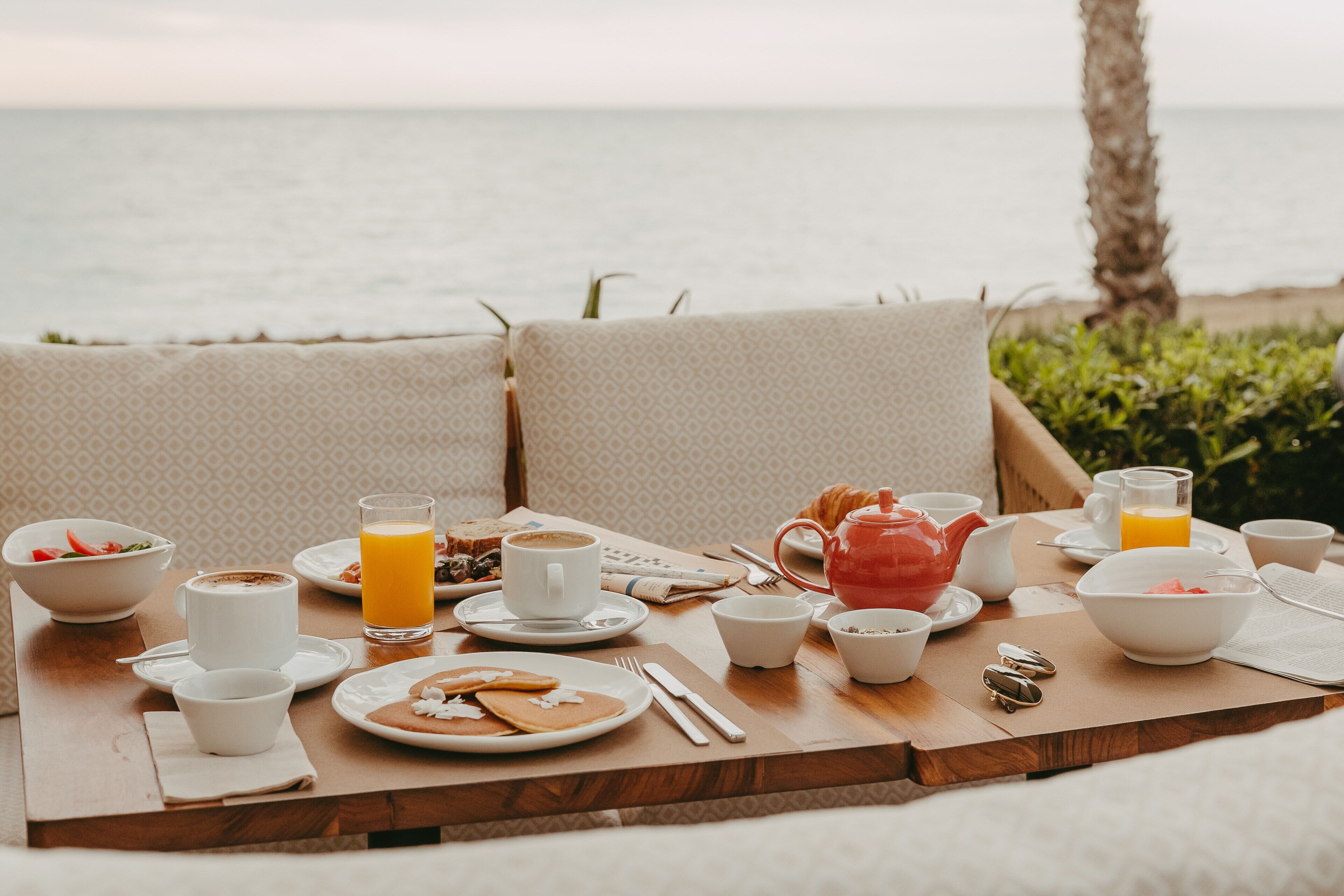 A breakfast setup on a wooden table with a view of the sea, including plates of food, cups, and a teapot.