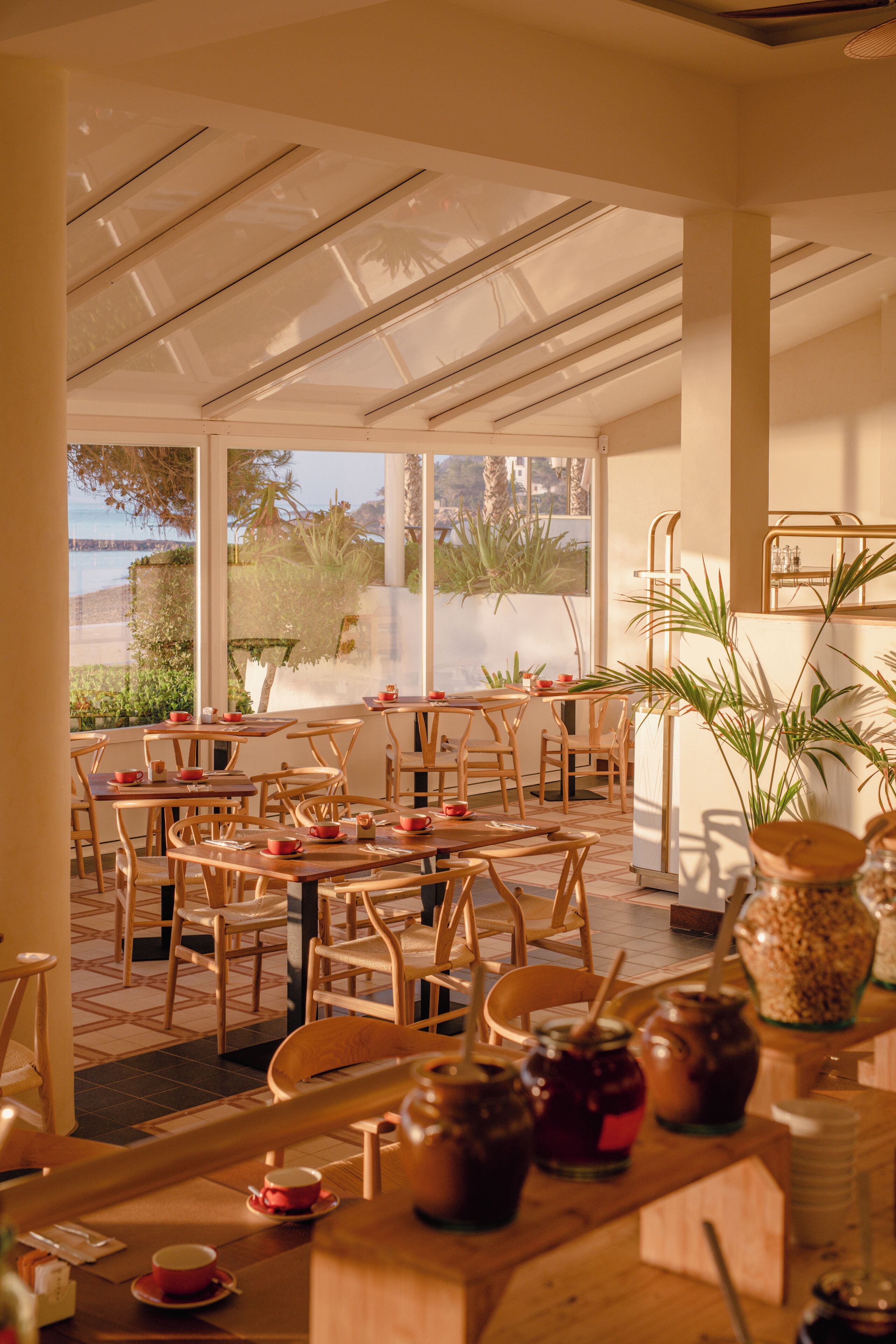 An indoor dining area with wooden tables and chairs, large windows with a view of the outdoors, and various jars and containers on the tables.