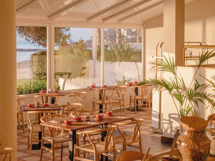 An indoor dining area with wooden tables and chairs, large windows with a view of the outdoors, and various jars and containers on the tables.