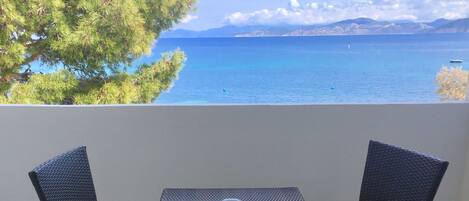 A balcony with a table and two chairs, overlooking a body of water and distant mountains.