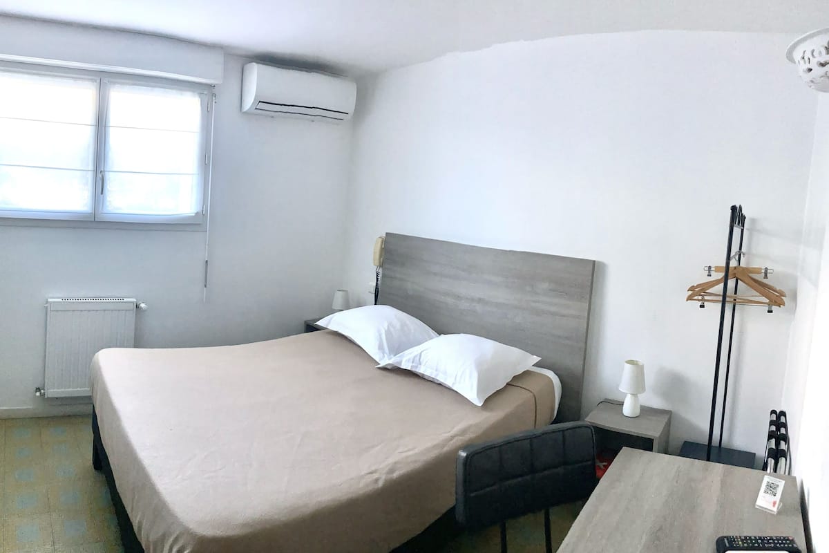 A hotel room with a bed, a desk, a chair, a radiator, an air conditioning unit, and a window.