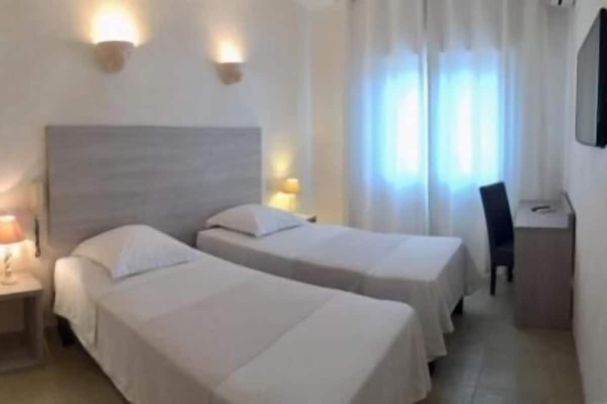 A hotel room with two single beds, a nightstand with a lamp, a desk with a chair, and a TV mounted on the wall.