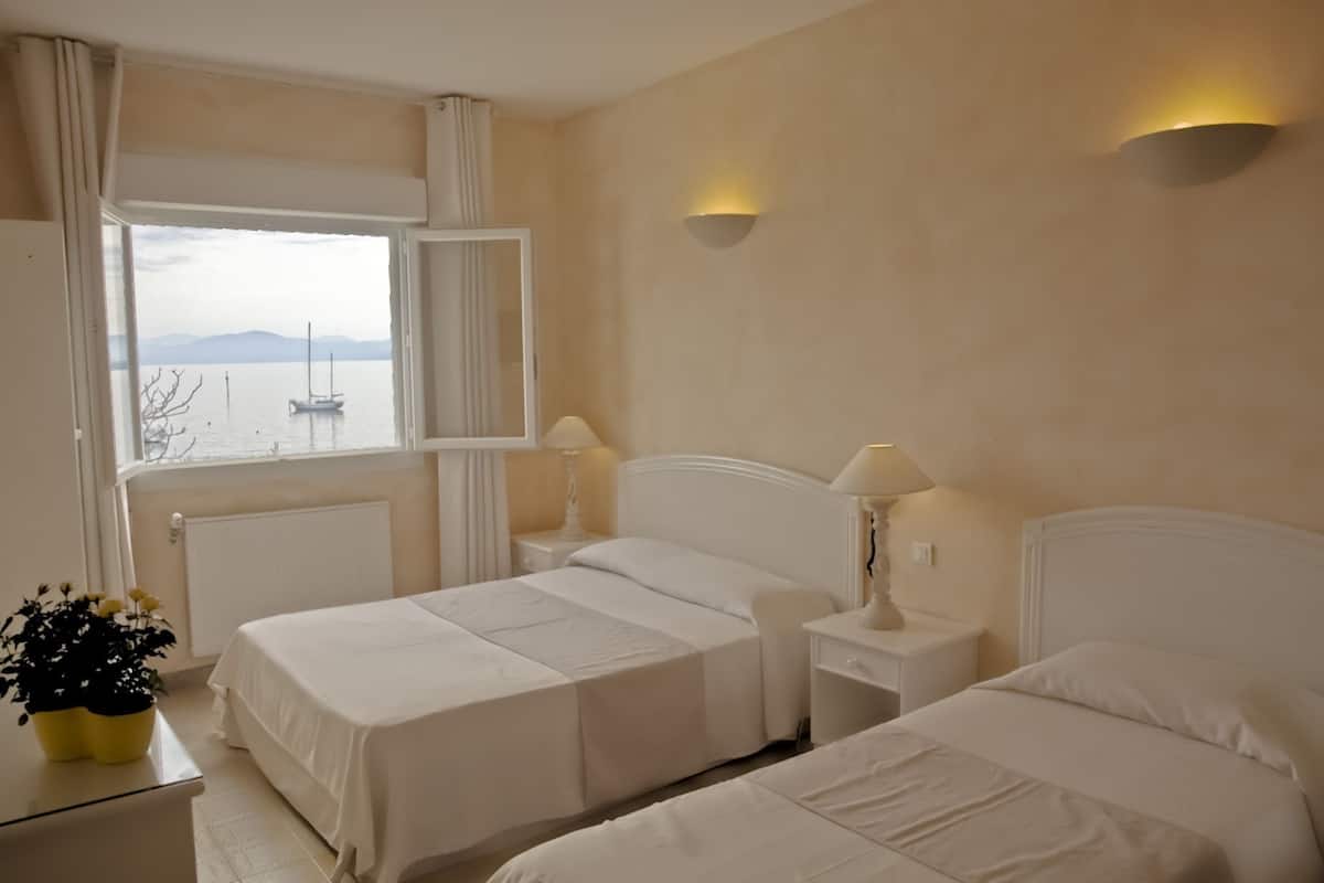 A hotel room with two beds, a window with a view of a body of water and mountains, and a potted plant on a nightstand.
