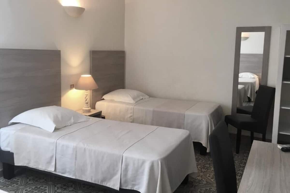 A hotel room with two single beds, a desk, a chair, a lamp, and a mirror.