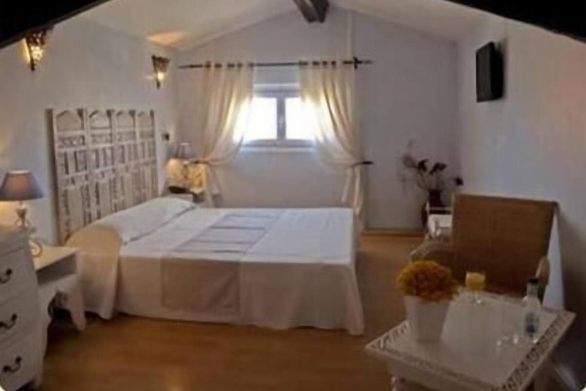 A bedroom with a large bed, a bedside table, a chair, and a small table with a vase.