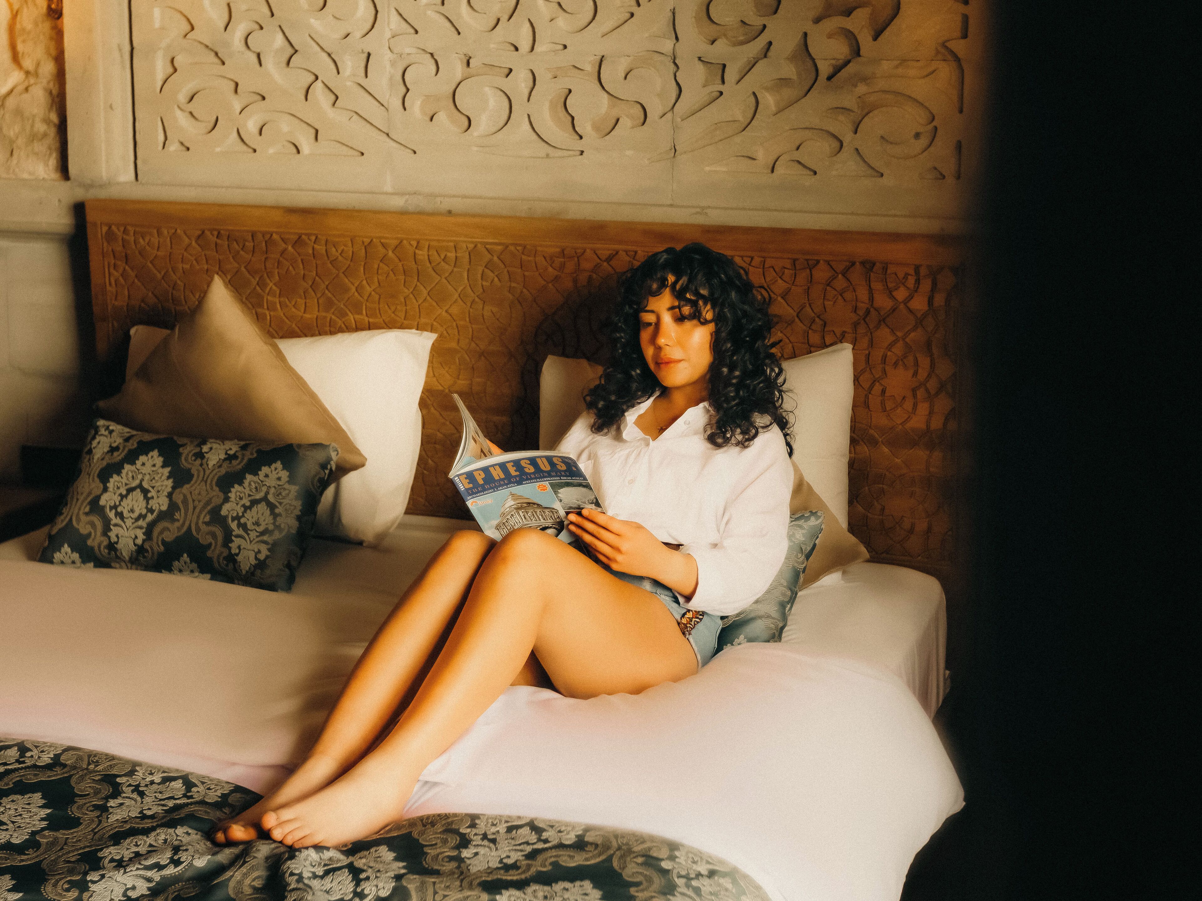 Una persona sentada en una cama con un libro, almohadas y una colcha estampada.