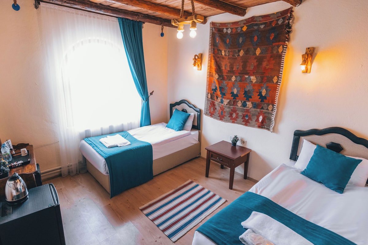 Habitación de hotel con dos camas, techo de madera, ventana con cortinas, lámpara de pared y una mesita con una planta.