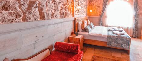 A room with a stone wall, a bed with a floral bedspread, a red upholstered bench, and a small wooden nightstand with a lamp.