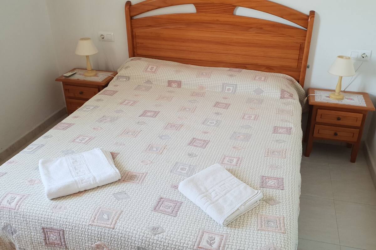 A neatly made bed with a patterned bedspread and two folded towels on it. There are two bedside tables with lamps and a small drawer.