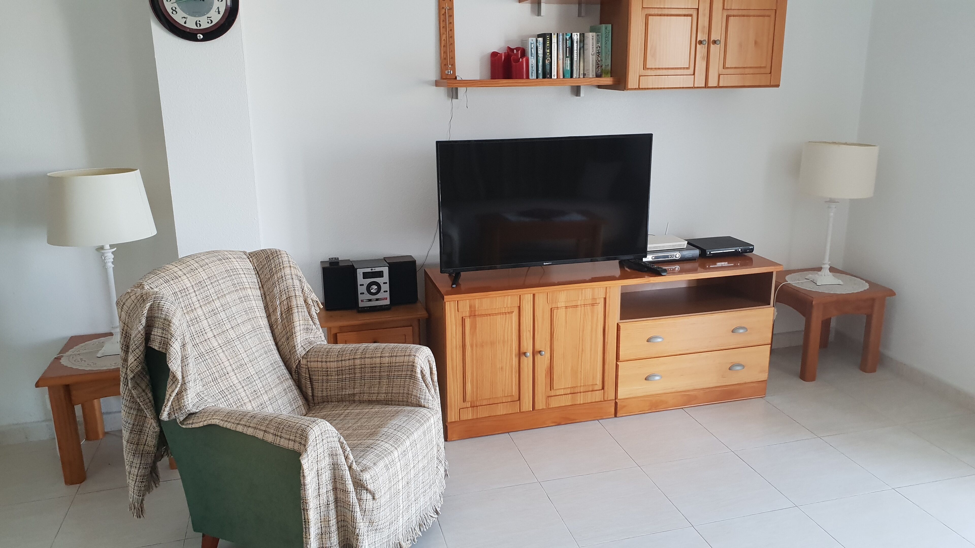 Una sala de estar con un televisor sobre un mueble de madera, un sillón verde, una mesita con una radio y una estantería con libros y objetos decorativos.