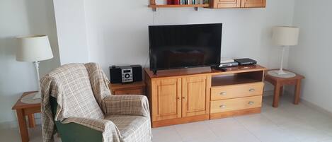 Una sala de estar con un televisor sobre un mueble de madera, un sillón verde, una mesita con una radio y una estantería con libros y objetos decorativos.