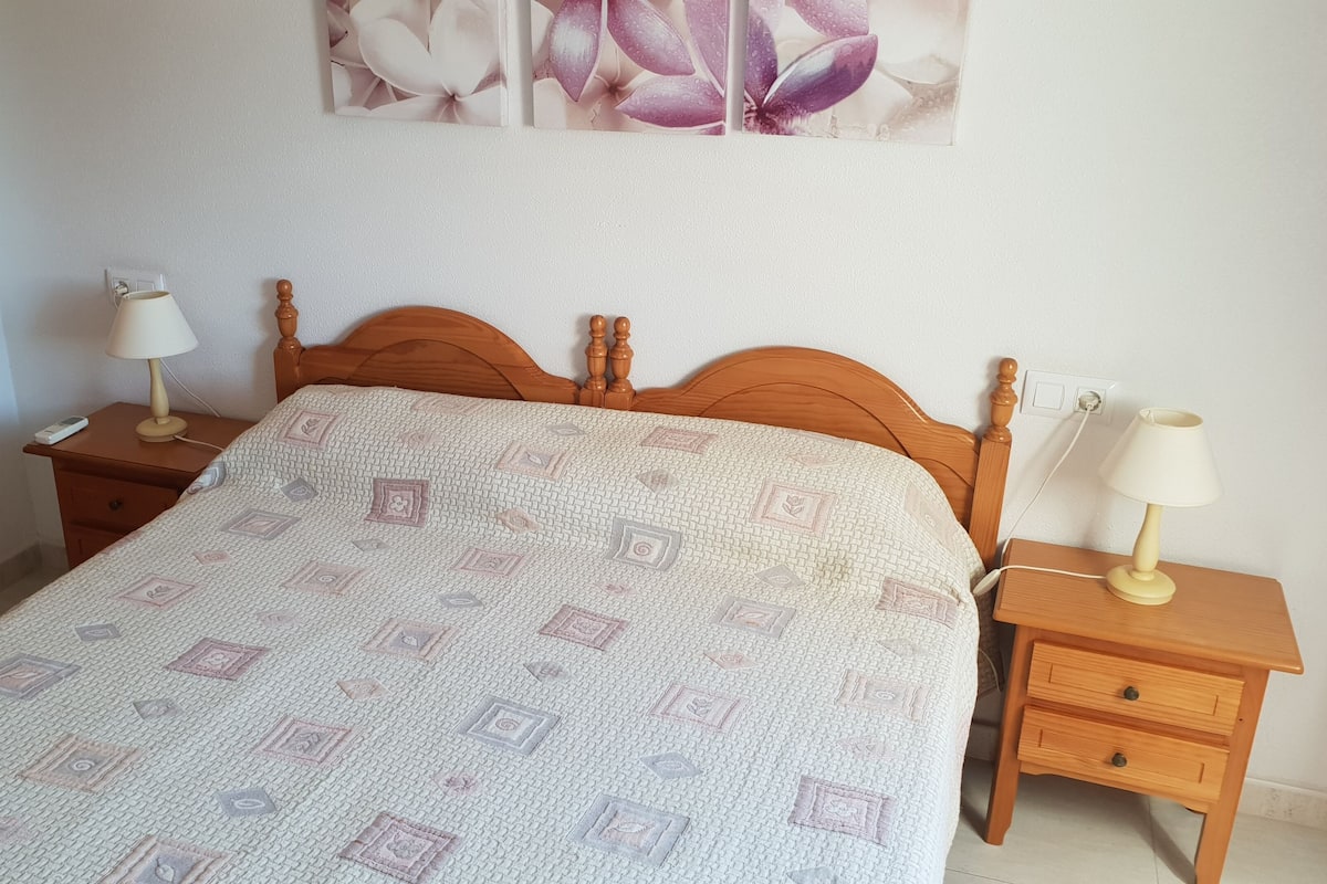 A neatly made bed with a patterned quilt, two bedside tables with lamps, and a wall-mounted power outlet.